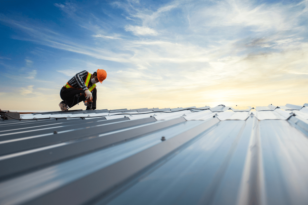 spring roofing upgrade. metal roofing in Walker, LA at Walker metals. A roof worker in safety gear installing a metal roof at sunset, using a power drill while secured with a harness.